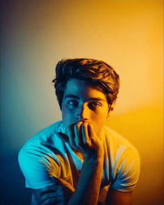 a young man with his hand on his chin looking at the camera while sitting in front of a wall