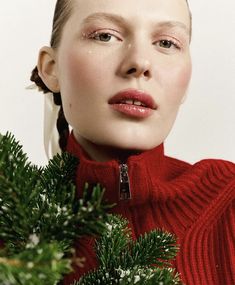 a woman wearing a red turtle neck sweater with braids on her head and green branches in front of her face