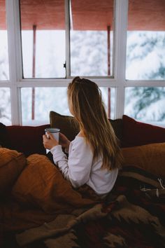 a woman sitting on a couch holding a cup in her hand and looking out the window