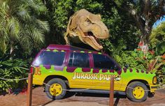 a large dinosaur statue sitting on the back of a van in front of trees and bushes