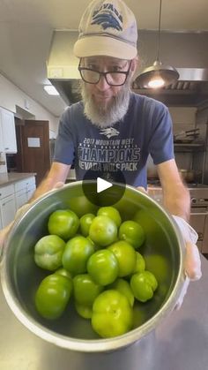 a man is holding a bowl full of green tomatoes in the kitchen, while wearing glasses and a hat