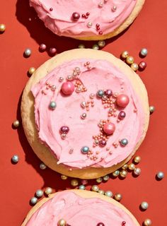 three cookies with pink frosting and sprinkles on red table top next to each other