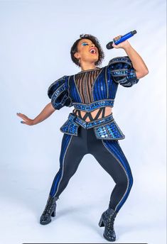 a woman in a blue and black outfit holding a microphone up to her face while standing on a white background