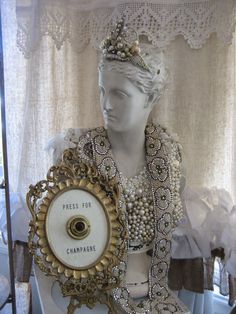 a white mannequin wearing pearls and a tiara next to a plaque that reads press for champagne