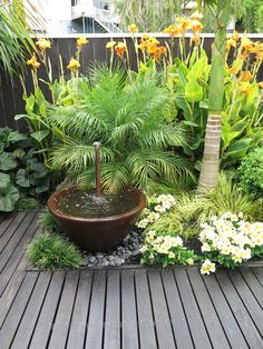 a water fountain surrounded by plants and flowers on a wooden deck in a garden area