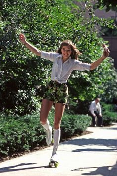 a young woman riding a skateboard down a sidewalk next to bushes and trees with her arms in the air