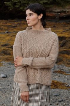 a woman standing in front of some rocks wearing a brown sweater and plaid skirt with her arms crossed