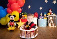 a baby boy sitting in a mickey mouse chair with balloons and other decorations behind him