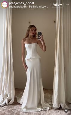 a woman in a white dress taking a selfie with her cell phone while standing next to curtains