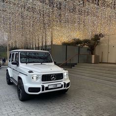 a white mercedes g - class is parked in front of some stairs with lights hanging from the ceiling