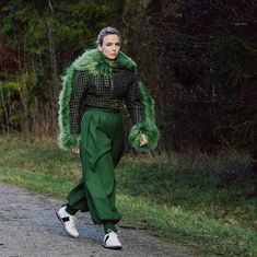 a woman walking down a road wearing green pants and a fur stole around her neck