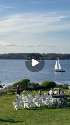 people are sitting on lawn chairs near the water and sailboats in the distance,