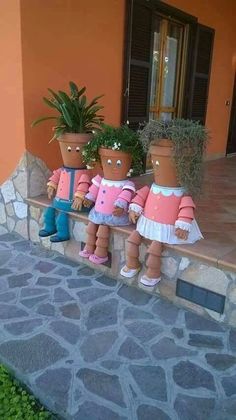 three little dolls sitting on the ledge of a stone wall with potted plants in front of them