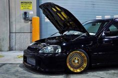 a black car with gold rims is parked in front of a garage door and it's hood open