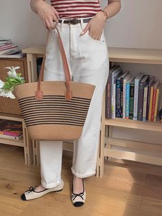 a woman in white pants holding a brown and black striped tote bag while standing next to a book shelf