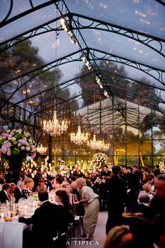 a large group of people sitting at tables in a room with chandeliers hanging from the ceiling