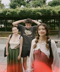 three young people standing next to each other in front of some trees and bushes on the sidewalk