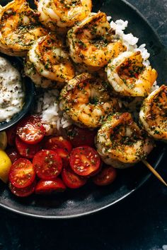 shrimp skewers with rice, tomatoes and lemon on a plate next to a bowl of sauce