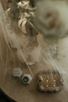 wedding shoes and jewelry sitting on a table with veil over it's head, in front of a white rose