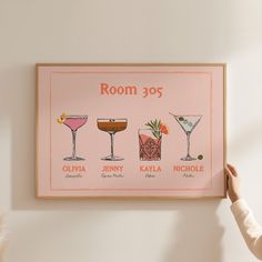 a woman holding up a framed poster with different cocktails on it's wall