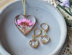 This beautiful handmade botanical jewelry set features a golden heart shaped pendant and earrings, with a purple Daisy and white Queen Anne's Lace flowers, sprouting from a reflective bed of crushed glass. Pressed Flowers that will never die,  preserved forever in crystal clear resin, with a beautiful domed finish.Flower Name & Meaning-  Daisy- new beginnings, rebirth, love. Queen Anne's Lace- safety, beauty, attract loveBezels/ Pendant Frame-  Heart shaped golden frame -1.2" wide x 1.3" tall -A Handmade Rose Gold Heart Pendant Jewelry, Mother's Day Flower Shaped Jewelry With Matching Earrings, Handmade Heart-shaped Jewelry As A Gift For Her, Mother's Day Flower Shaped Jewelry With Heart Charm, Mother's Day Flower Jewelry With Heart Charm, Mother's Day Jewelry With Heart Charm And Flower Shape, Nature-inspired Heart Shaped Jewelry Gift, Handmade Heart-shaped Jewelry For Mother's Day, Delicate Valentine's Day Jewelry With Matching Earrings