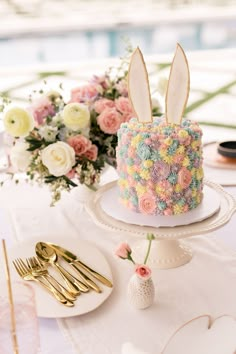 a cake decorated with flowers and bunny ears is on a table next to gold utensils