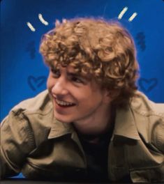 a man with curly hair is smiling and looking at the camera while sitting in front of a blue background