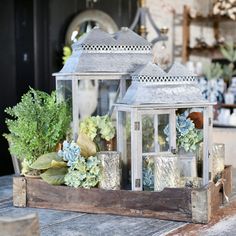 two white lanterns sitting on top of a wooden tray filled with flowers and candles next to potted plants