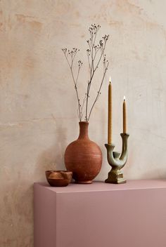 a vase with two candles and a bowl on a pink shelf in front of a wall