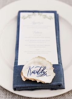 the place setting is set with an oyster shell and blue linen napkins, which are adorned with handwritten calligraphy