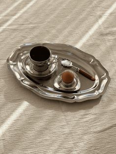 a silver tray with an egg, spoon and cup on it sitting on a bed