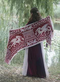 a woman holding a large red and white scarf in front of a lake with willow trees