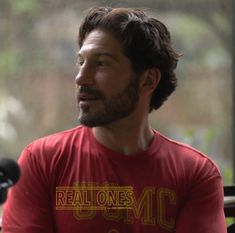 a man with long hair and beard wearing a red t - shirt sitting in front of a window