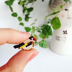 a hand holding a tiny beaded bird on it's finger next to a potted plant