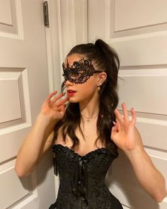 a woman in a black corset and mask poses with her hands behind her head