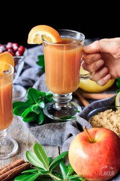 two glasses filled with orange juice on top of a table next to apples and cinnamons