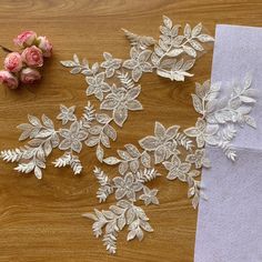 two pieces of white lace next to pink flowers on a wooden table with a piece of paper