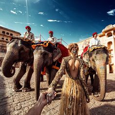 a woman in a gold dress standing next to two elephants with people riding on them