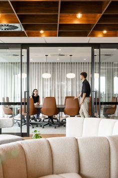 two people are standing in an office with glass walls and wooden ceilinging, while another person is sitting at a table