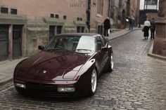 a maroon sports car parked on the side of a cobblestone street in an old european city