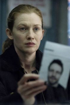 a woman looking at her cell phone while holding an envelope with a man in the background