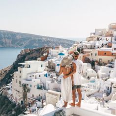 two people standing on the edge of a cliff with white buildings and blue water in the background