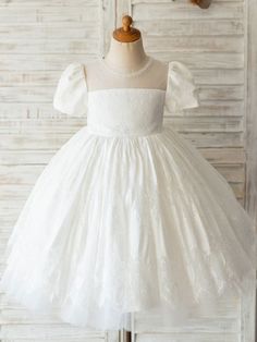 a white flower girl dress on a mannequin dummy with flowers in the background