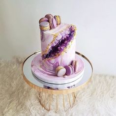 a pink cake with purple icing and gold decorations on a stand in front of a white wall