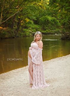 a pregnant woman in a pink dress standing on the beach near a river and trees