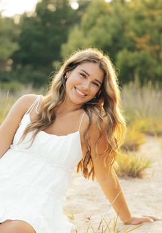 a beautiful young woman sitting on top of a sandy beach next to green grass and trees