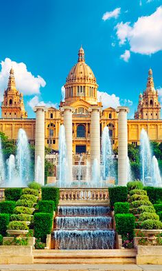 an ornate building with fountains in the foreground and blue sky above it, surrounded by greenery