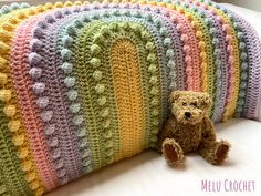 a teddy bear sitting next to a multicolored crocheted pillow on a bed