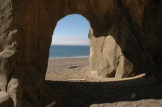 an arch in the side of a rock formation near the beach with water behind it