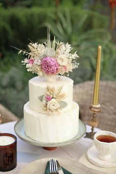 a white wedding cake with pink flowers on top sitting on a table next to a cup and saucer